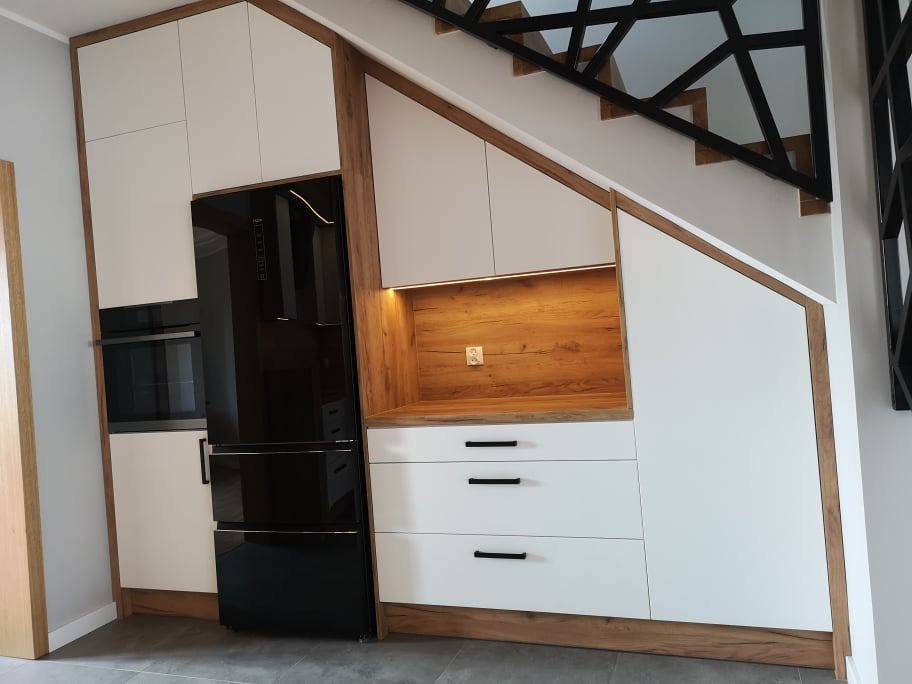 Białe szafki kuchenne z drewnianym obramowaniem, zabudowa pod schodami, czarna lodówka i piekarnik w zabudowie, szare płytki podłogowe.
