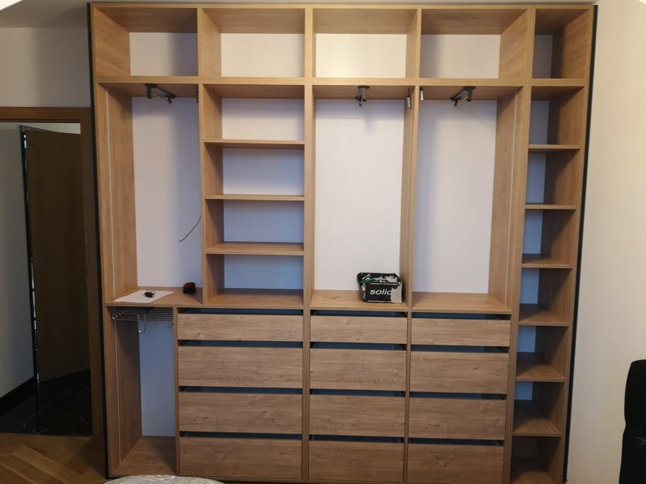 Wnętrze szafy z jasnego drewna w trakcie montażu: widoczne półki, szuflady, drążki na ubrania, pudełko narzędzi na blacie, częściowo zamontowana i otwarta.