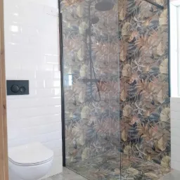 Nowoczesna kabina prysznicowa z motywem liści na płytkach, sąsiadująca z białą toaletą podwieszaną i błyszczącymi, białymi płytkami ściennymi typu metro.