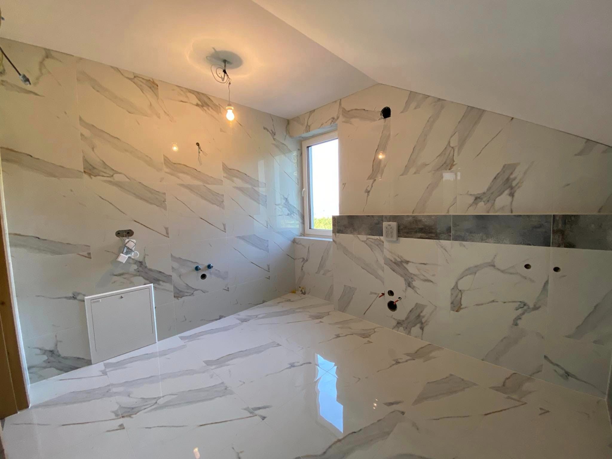 Łazienka w trakcie wykańczania z marmurowymi płytkami na ścianach i podłodze, widoczne otwory na armaturę, sufit pod skosem i okno z widokiem na zieleń.