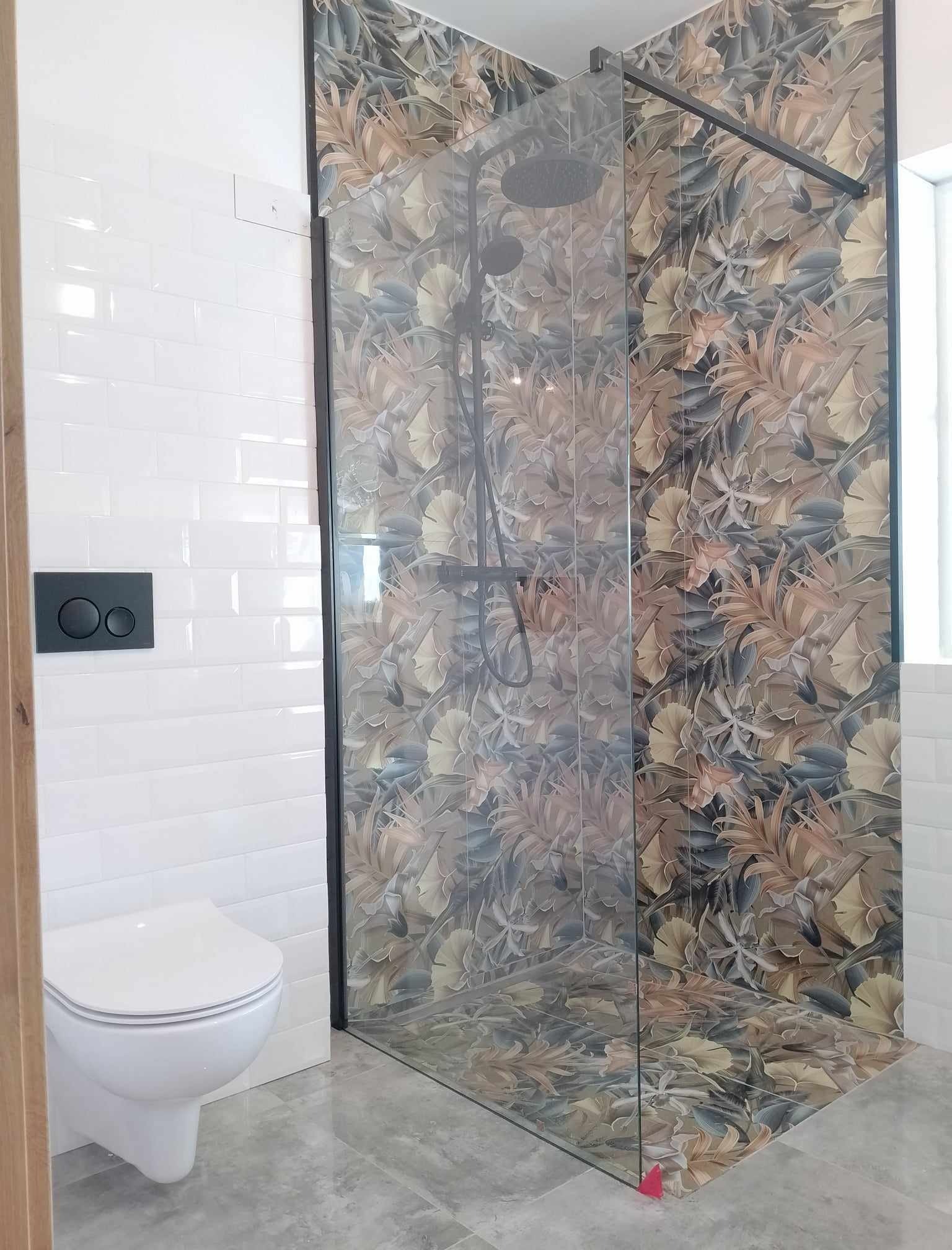 Nowoczesna kabina prysznicowa z motywem liści na płytkach, sąsiadująca z białą toaletą podwieszaną i błyszczącymi, białymi płytkami ściennymi typu metro.