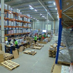 Logistic Space Sp zo.o. - Wnętrze magazynu z wysokimi regałami wypełnionymi zapakowanymi towarami, pracownicy w zielonych koszulkach przy biurkach, palety z kartonami i gotowe paczki.