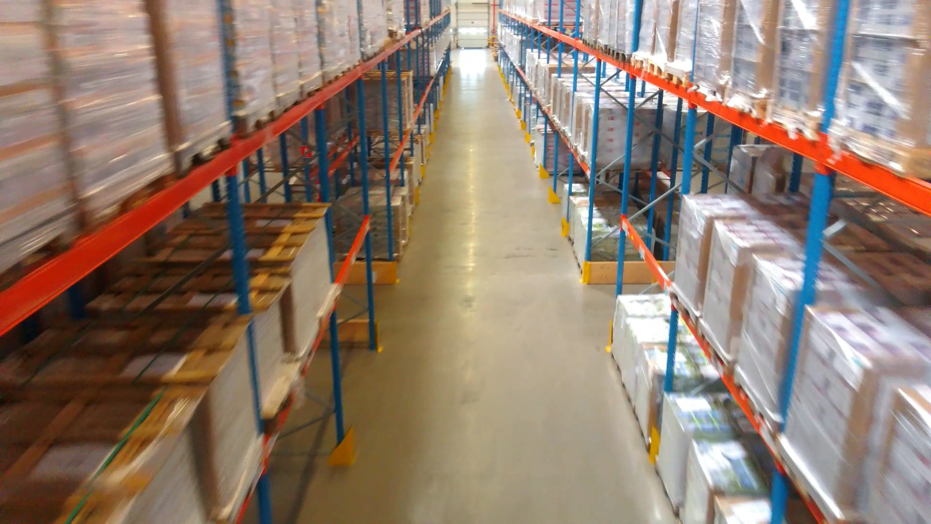 Perspektywa długiego korytarza magazynowego z regałami wypełnionymi paletami, zabezpieczonymi folią stretch, z widocznymi niebieskimi i czerwonymi elementami konstrukcyjnymi regałów.