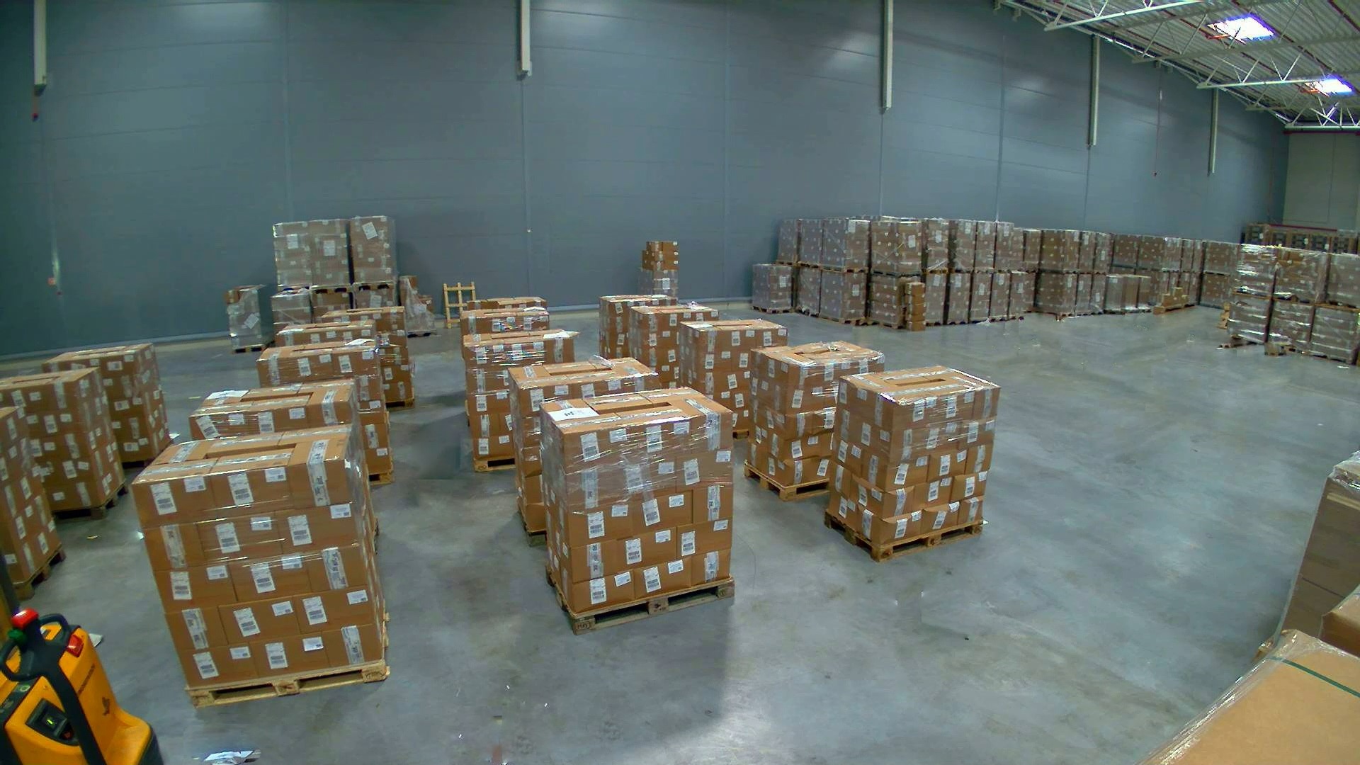 Wnętrze magazynu z paletami kartonowych pudeł owiniętych folią, ustawionych w rzędach, widoczny wózek widłowy z prawej strony kadru.