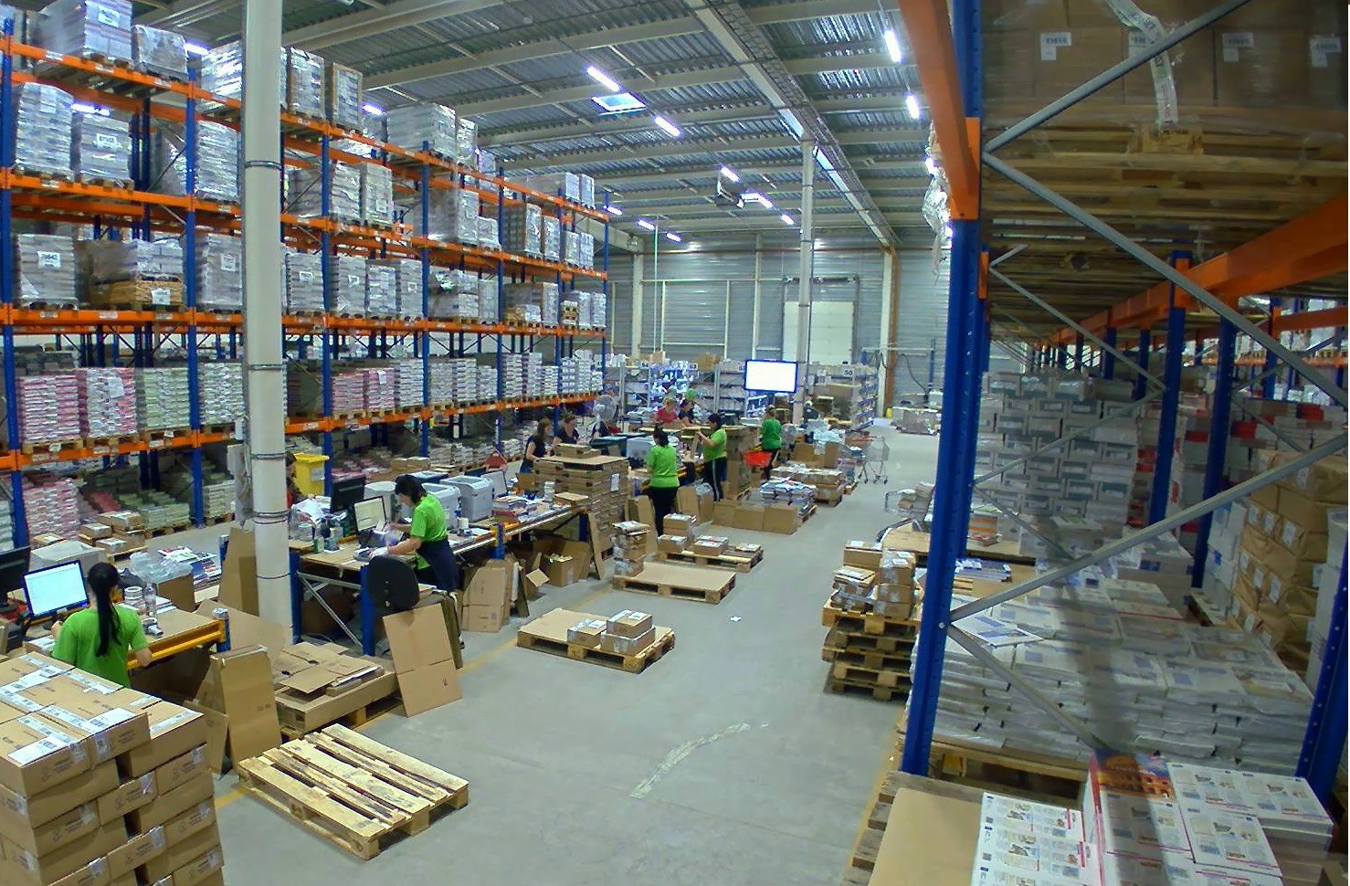 Wnętrze magazynu z wysokimi regałami wypełnionymi zapakowanymi towarami, pracownicy w zielonych koszulkach przy biurkach, palety z kartonami i gotowe paczki.