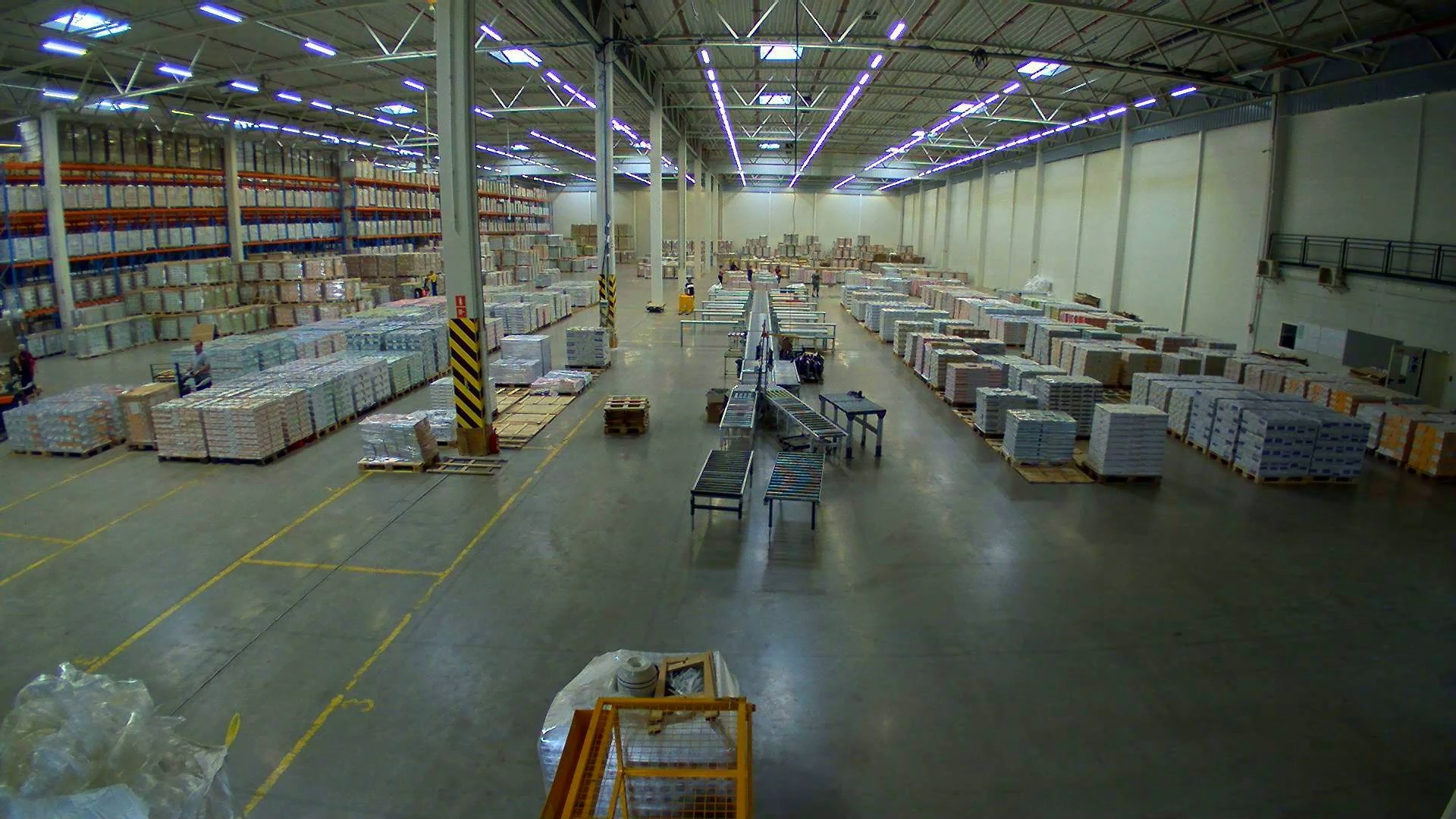 Widok z góry na przestronny magazyn z regałami wypełnionymi zapakowanymi towarami na paletach, z widocznymi wózkami widłowymi i systemem przenośników rolkowych.