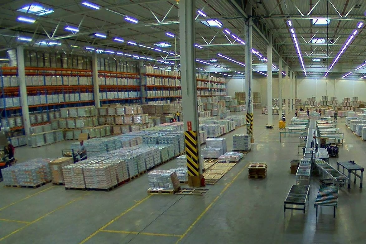 Widok z góry na duży magazyn z regałami pełnymi zapakowanych towarów, pracownicy obsługują wózki widłowe i przenośniki taśmowe.