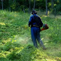 Pracownik w niebieskim kombinezonie i słuchawkach tnący wysoką trawę na zboczu wzgórza za pomocą pomarańczowej podkaszarki Husqvarna, w tle widoczne drzewa.