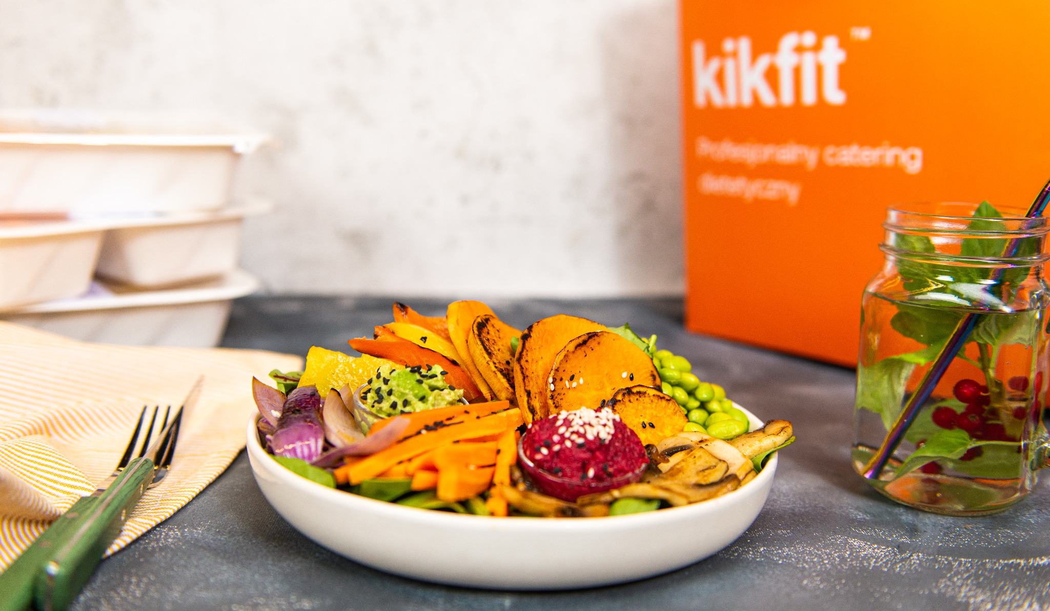 Kompozycja warzyw grillowanych i gotowanych w białej misce, obok napój w słoiku ze słomką i pudełka z logo Kikfit w tle.