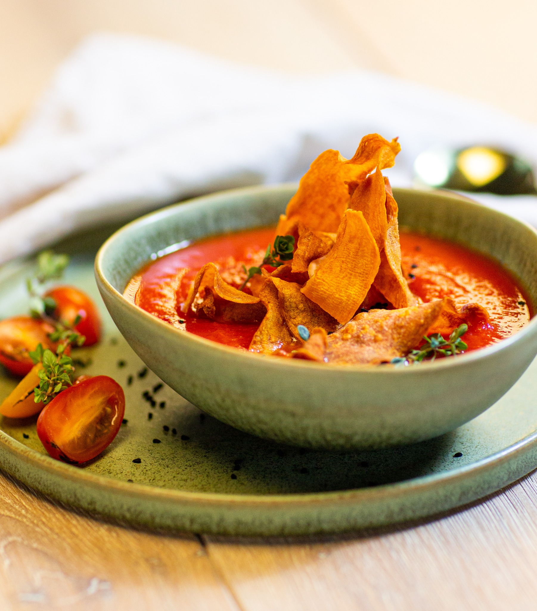 Krem z pomidorów z chipsami z batatów i świeżymi ziołami, podany w turkusowej miseczce na pasującym talerzu, z dekoracją z przekrojonych pomidorków koktajlowych i czarnych ziaren.