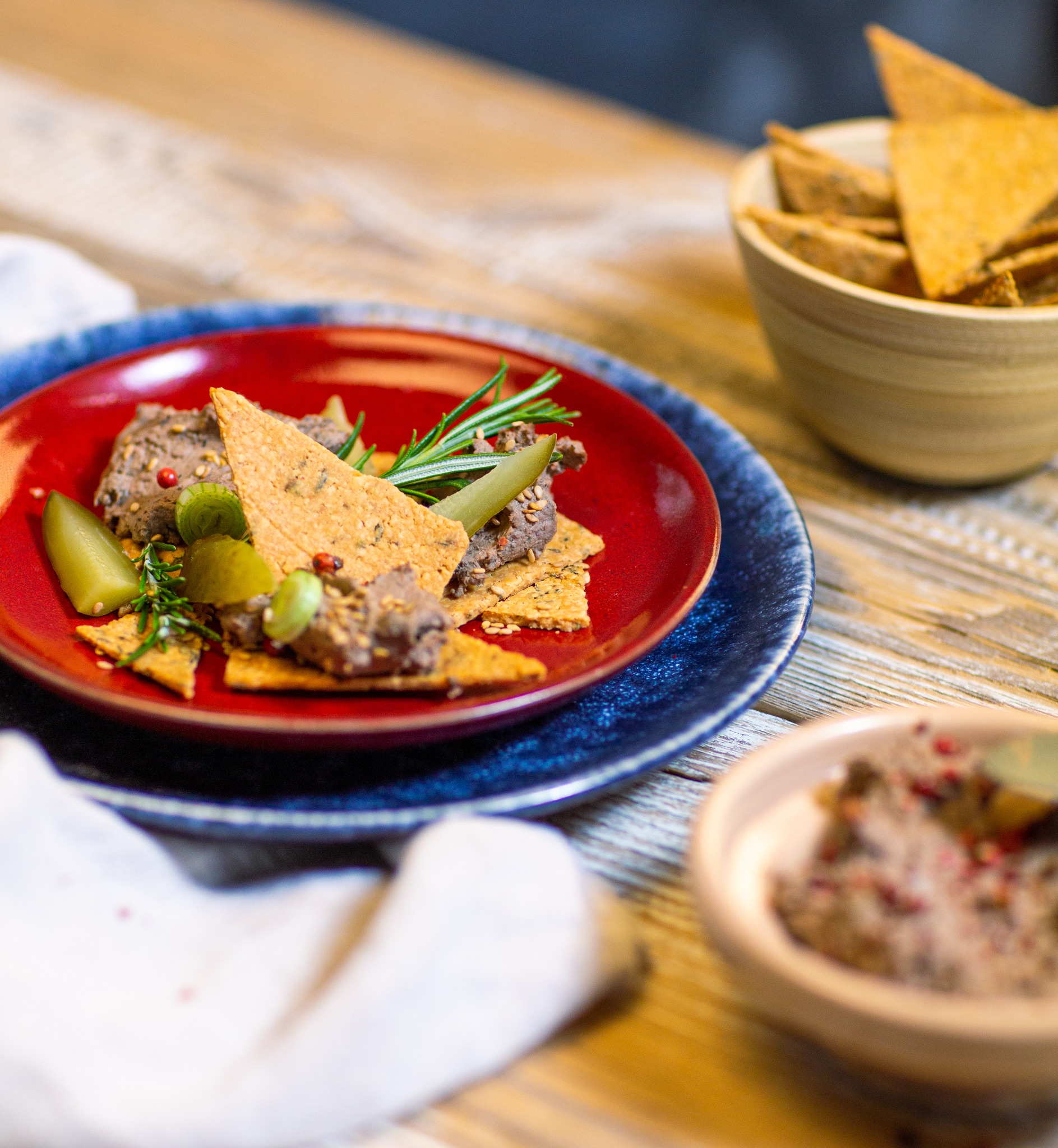 Kompozycja z pasztetem na krakersach, udekorowana korniszonami i gałązką rozmarynu na czerwonym talerzu, obok miseczka z krakersami i druga z pasztetem, na drewnianym stole.