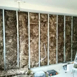 Ściana wewnętrzna z metalowym stelażem wypełnionym brązową wełną mineralną, częściowo zakryta, widoczna drabina i narzędzia na podłodze.