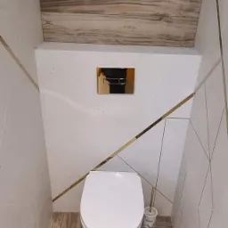 Nowoczesna toaleta w łazience wykończonej płytkami imitującymi drewno na podłodze i suficie, białe ściany z geometrycznym wzorem i złotymi paskami, złoty przycisk spłuczki.