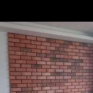 Ściana wyłożona dekoracyjną cegłą w odcieniach czerwieni i brązu, z widocznymi fugami. Nad cegłą biały pas sufitu i listwa przypodłogowa.