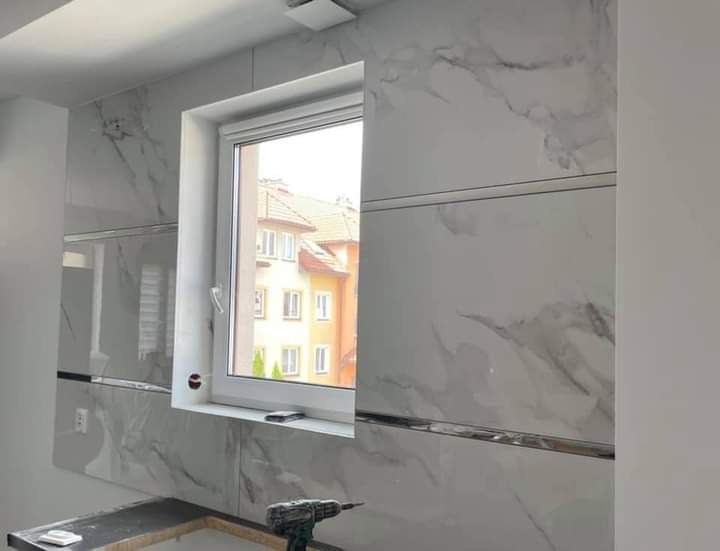 Ściana wyłożona białymi płytkami imitującymi marmur z metalowymi listwami, okno z widokiem na budynek mieszkalny oraz wiertarka akumulatorowa na blacie.