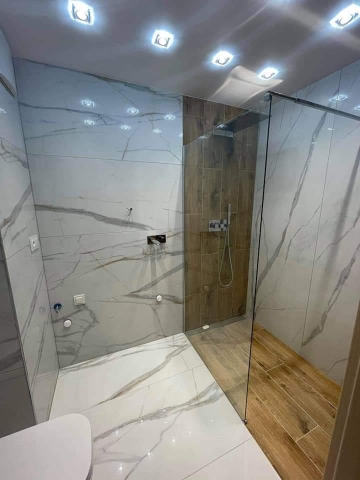 Nowoczesna łazienka z kabiną prysznicową typu walk-in, wykończona białymi płytkami imitującymi marmur oraz drewnopodobnymi płytkami w strefie prysznica, oświetlona punktowymi lampami sufitowymi.