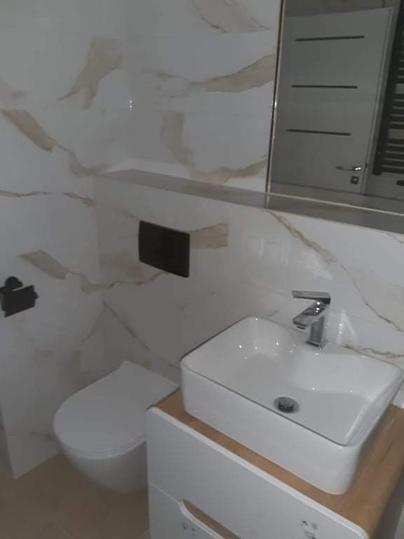 Nowoczesna łazienka z białymi marmurkowymi płytkami, wiszącą toaletą, prostokątną umywalką nablatową na szafce z drewnianym blatem i dużym lustrem z szafką. Minimalistyczny design.