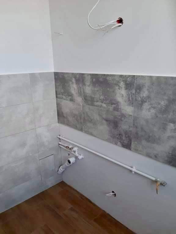 Ściana łazienki z częściowo ułożonymi szarymi płytkami imitującymi kamień, widoczne białe rury kanalizacyjne i wystające przewody elektryczne, drewnopodobna podłoga.