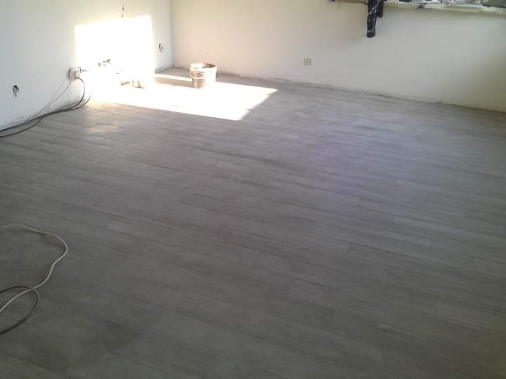 Ułożony parkiet w szarym odcieniu w pokoju z białymi ścianami, widoczne gniazdka elektryczne i wiadro w rogu, częściowo oświetlone wpadającym światłem słonecznym.