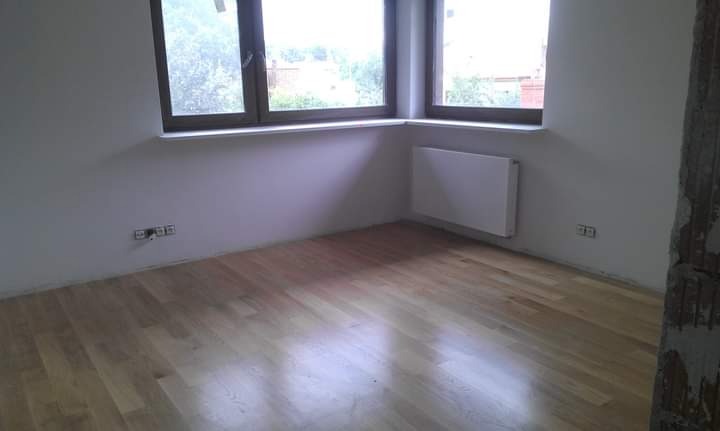 Jasny, świeżo ułożony parkiet w pustym pokoju z dwoma oknami i grzejnikiem na ścianie. Fragment ściany z widocznymi uszkodzeniami w prawym rogu.