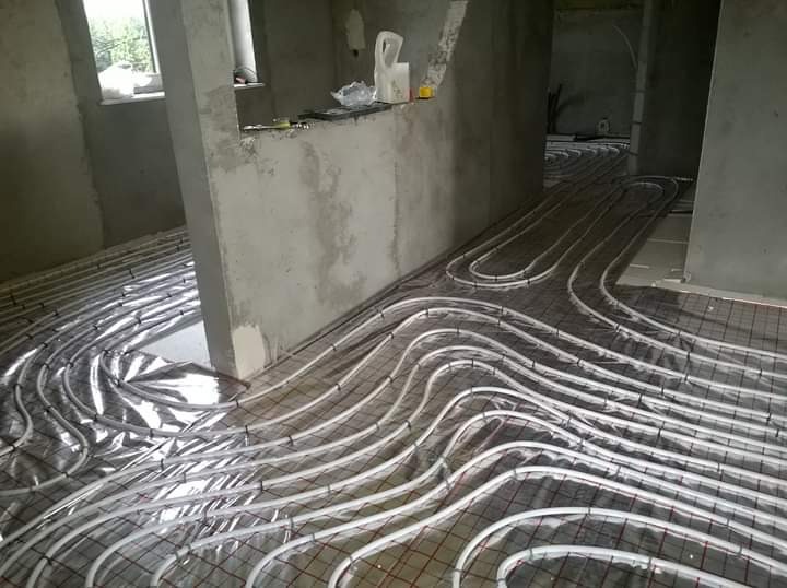 Rozłożone rury ogrzewania podłogowego na folii izolacyjnej z siatką, widoczne surowe ściany w trakcie remontu.