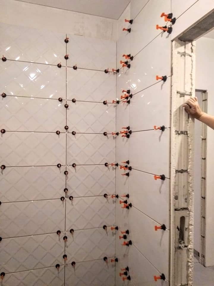 Ściana łazienki w trakcie układania białych płytek z użyciem systemu poziomowania, widoczna ręka przy ościeżnicy.