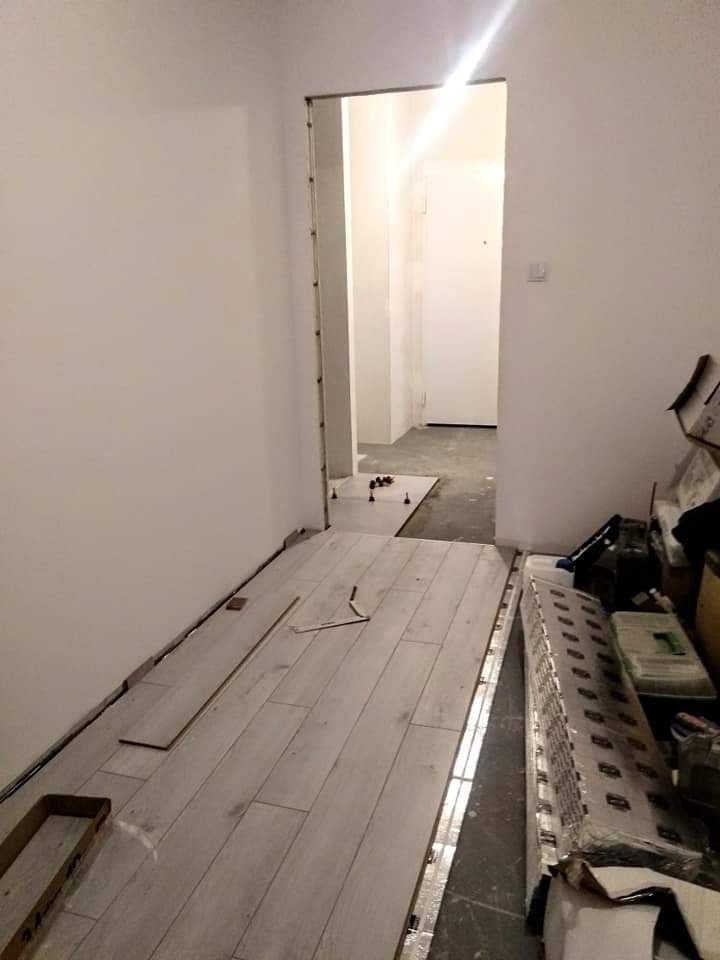 Układanie jasnych paneli podłogowych w korytarzu, widoczne narzędzia pomiarowe i paczki z panelami obok ściany, przygotowane do dalszego montażu.