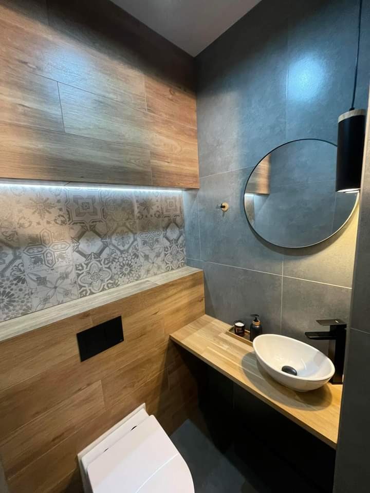 Nowoczesna łazienka z drewnianymi i szarymi płytkami, okrągłym lustrem, białą umywalką nablatową i czarną armaturą, oświetleniem LED nad dekoracyjnymi płytkami.