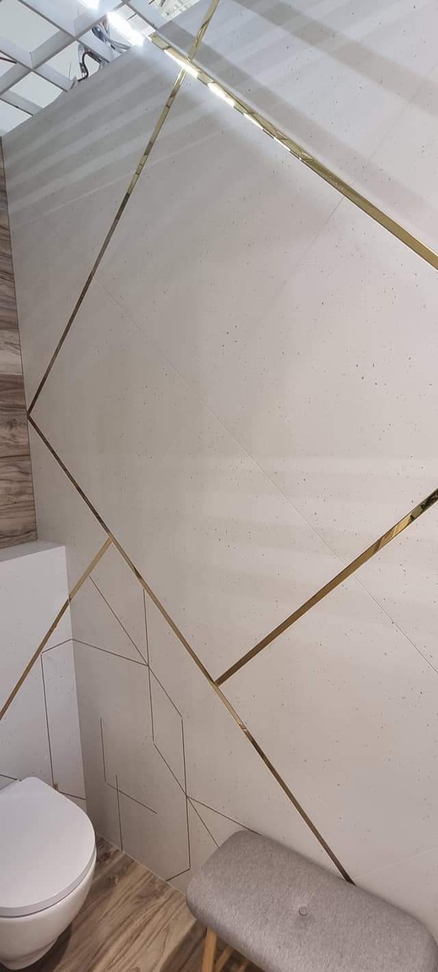 Białe płytki ścienne z geometrycznym wzorem podkreślonym złotymi listwami w wyremontowanej łazience, widoczny fragment toalety i szarego stołka.