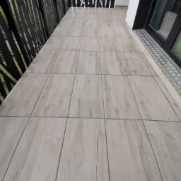 Wyłożony płytkami balkon z dekoracyjną, ażurową balustradą i odpływem wzdłuż okna balkonowego.