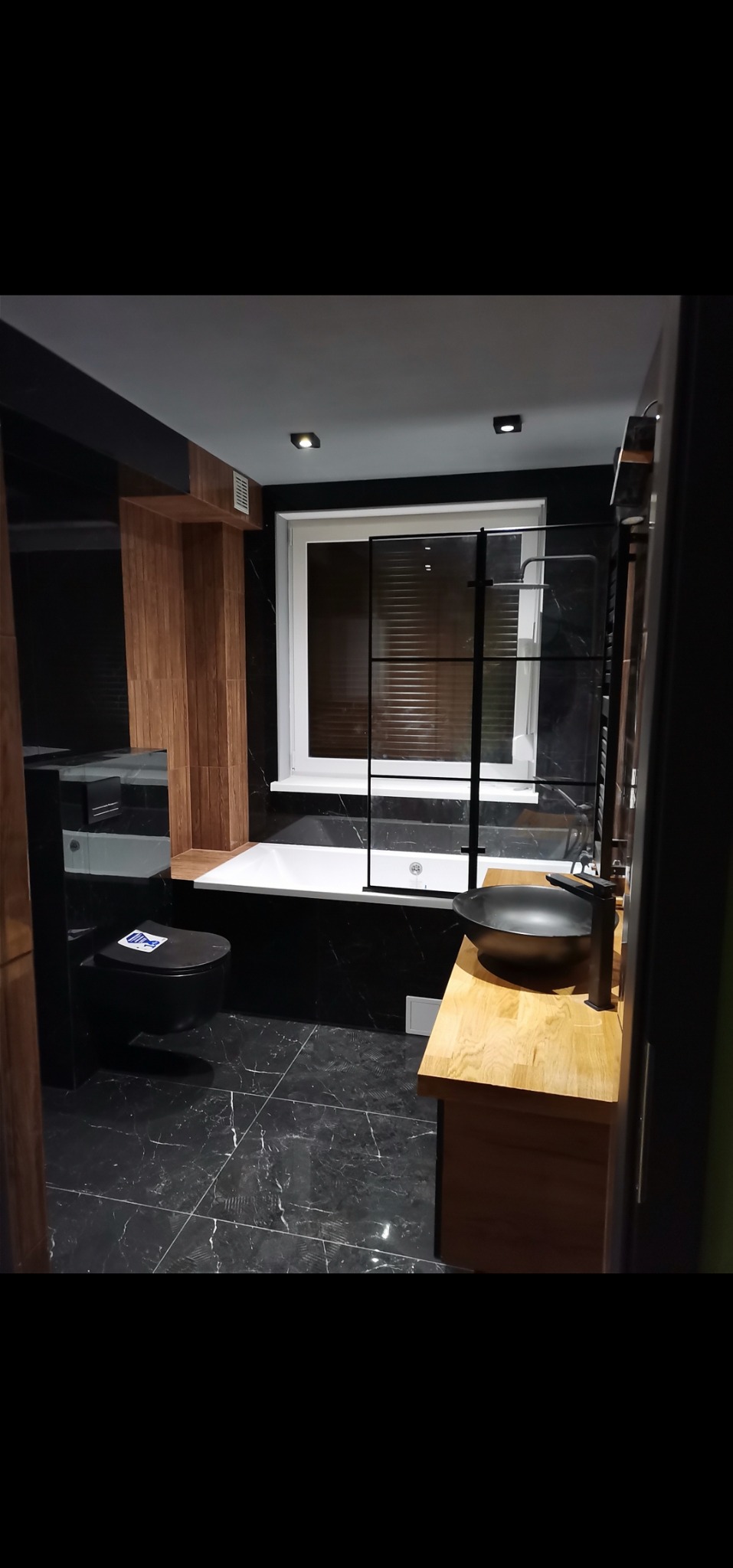 Elegancka łazienka w czerni i drewnie: czarna armatura, umywalka nablatowa i marmurowe płytki podłogowe, akcenty drewniane na ścianie i blacie, kabina prysznicowa typu walk-in z czarną ramą, okno...
