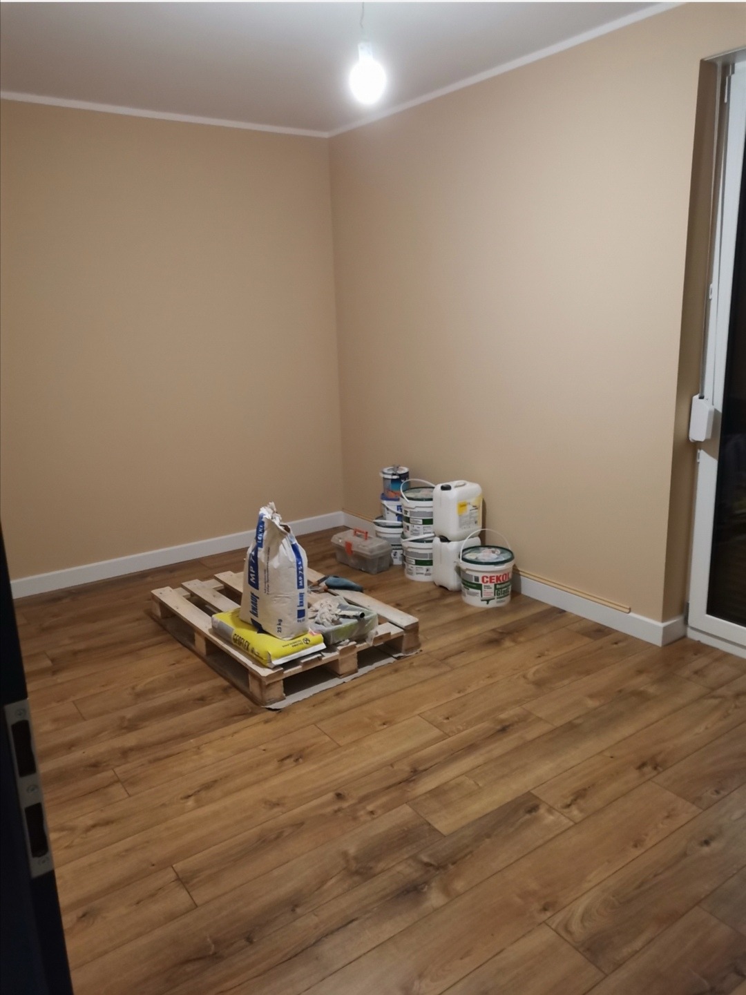 Pokój z beżowymi ścianami i brązowymi panelami podłogowymi, w rogu paleta z workiem zaprawy, obok puszki farb i narzędzia przygotowane do prac wykończeniowych.