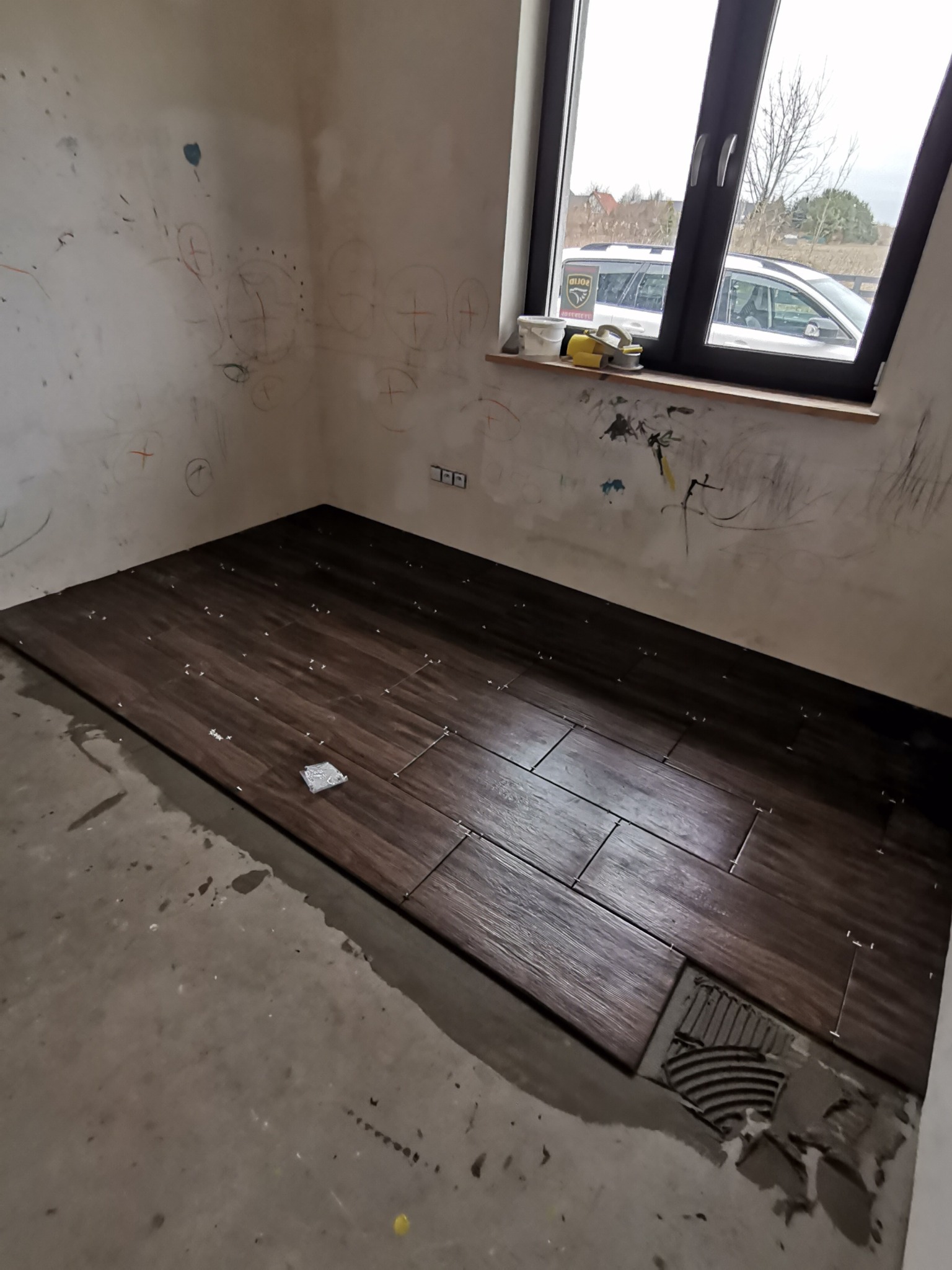 Układanie ciemnobrązowych płytek imitujących drewno w pokoju, widoczne nierówne ściany z zabrudzeniami i rysunkami, okno z widokiem na samochód i drzewa.