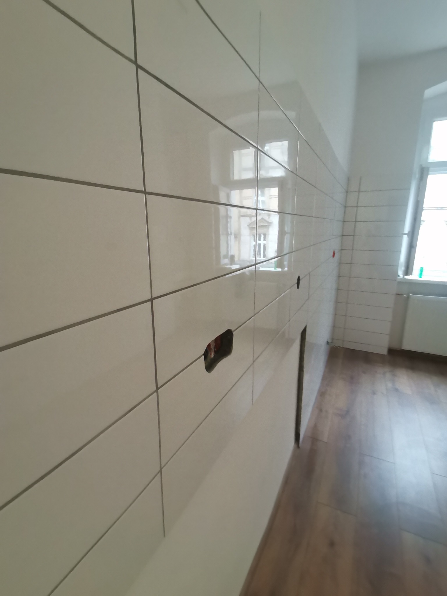 Świeżo ułożone, błyszczące, białe płytki ścienne z szarą fugą w łazience, widoczne otwory na gniazdka elektryczne i rury, na podłodze panele imitujące drewno, okno z widokiem na budynek.