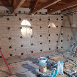 Ocieplenie ceglanej ściany poddasza z widocznymi kołkami mocującymi, przygotowane do dalszych prac wykończeniowych.