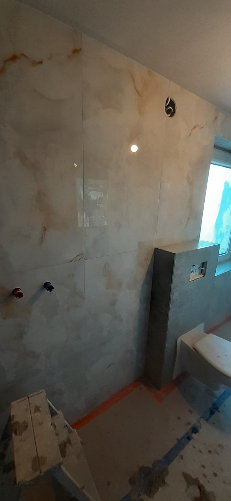 Ściana łazienki wyłożona płytkami imitującymi marmur, widoczne instalacje wodne i stelaż podtynkowy WC, w tle okno i drabina malarska.