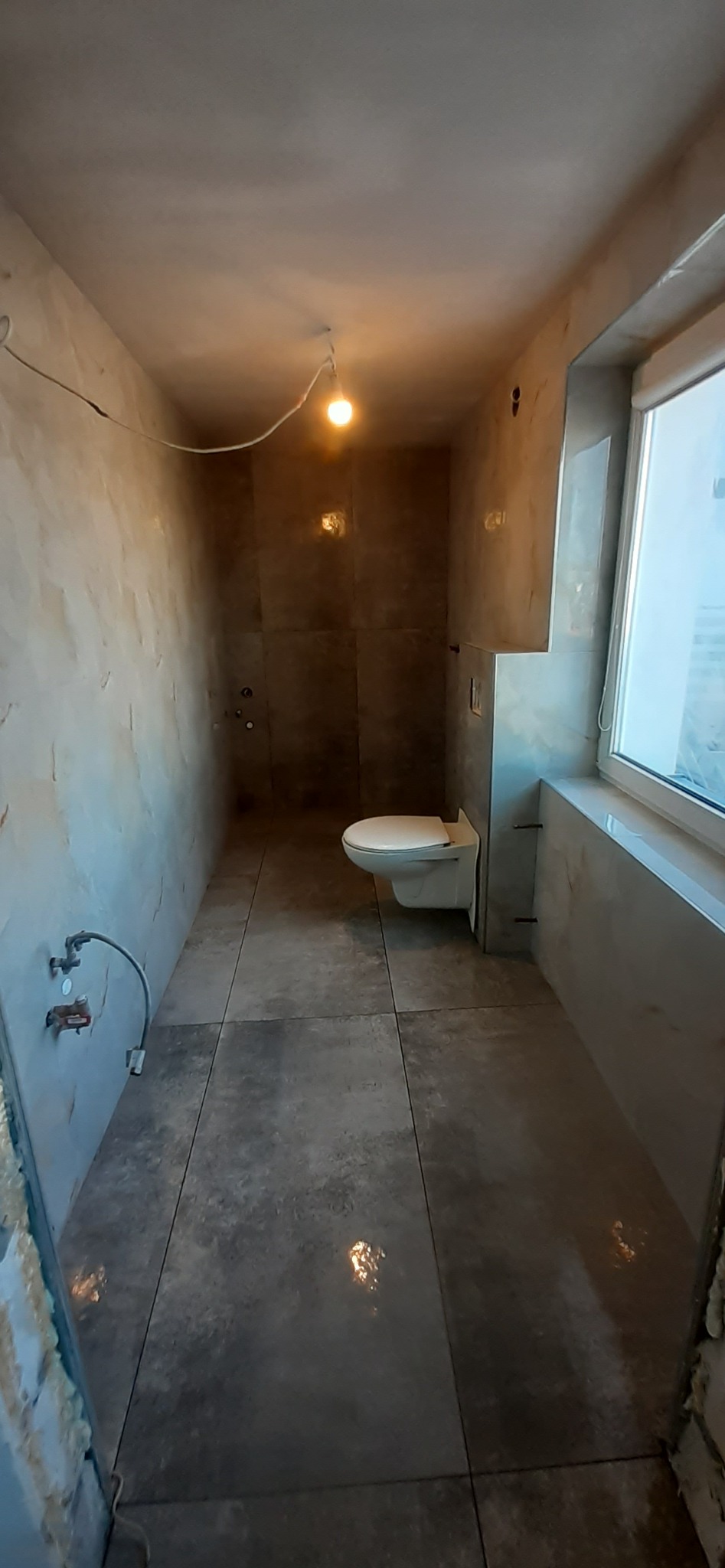 Wnętrze łazienki w trakcie remontu: widoczne płytki imitujące kamień na podłodze i ścianach, podwieszana toaleta oraz wystające przewody i niedokończone ściany.