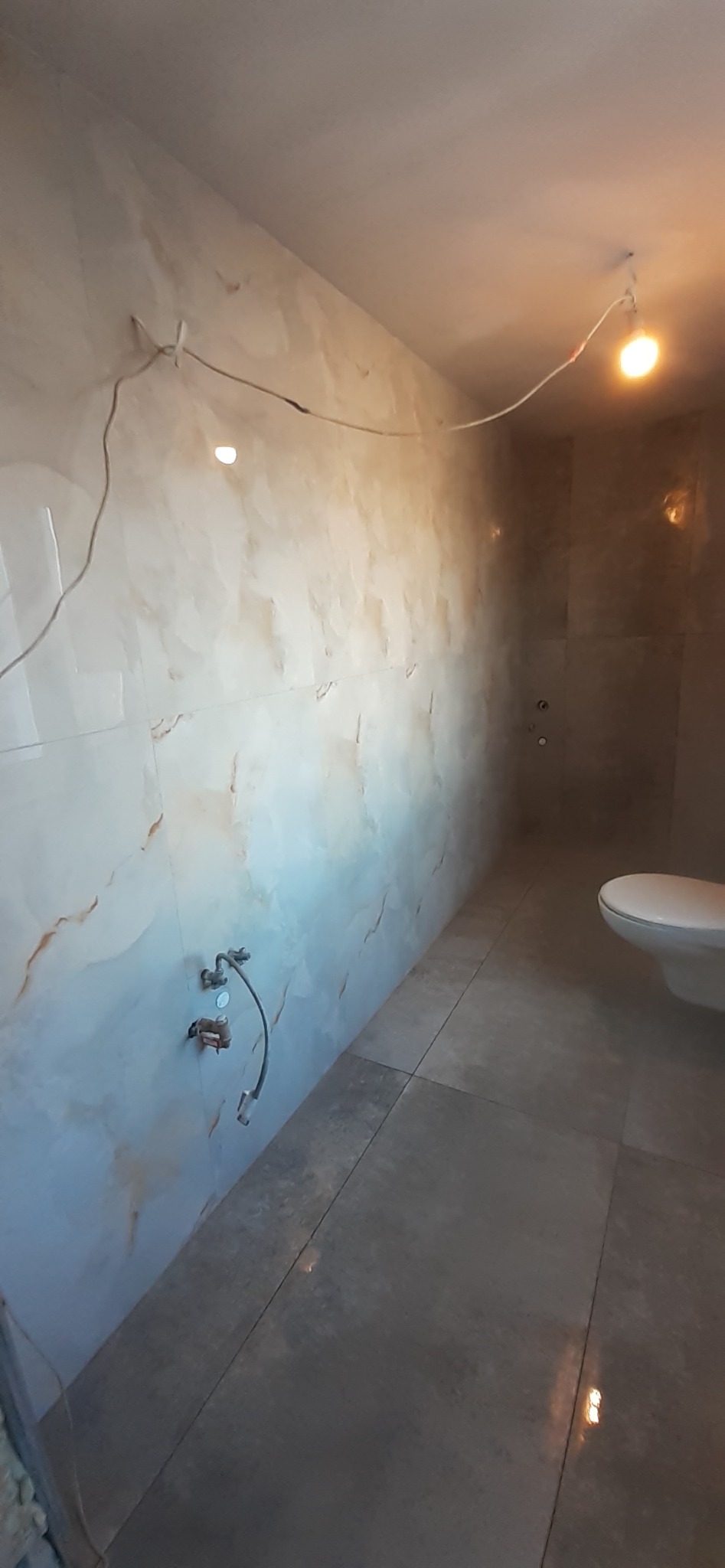 Wnętrze łazienki w trakcie prac wykończeniowych, widoczne płytki imitujące marmur na ścianach i podłodze, wystające kable i rury, biała toaleta w rogu.