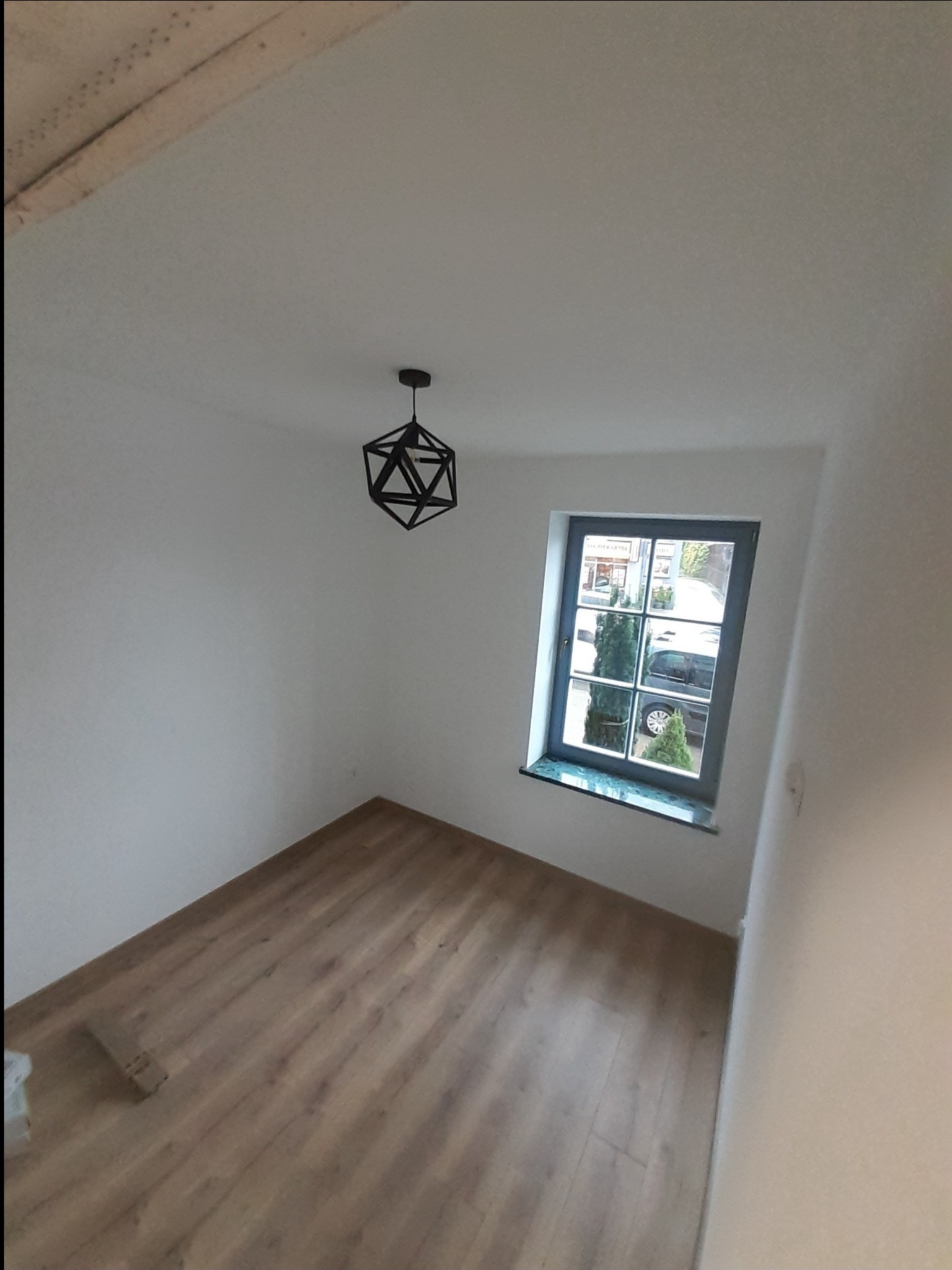 Pusty pokój po odświeżeniu: panele podłogowe imitujące drewno, białe ściany, okno z szarą ramą i marmurowym parapetem, oraz geometryczna lampa wisząca.