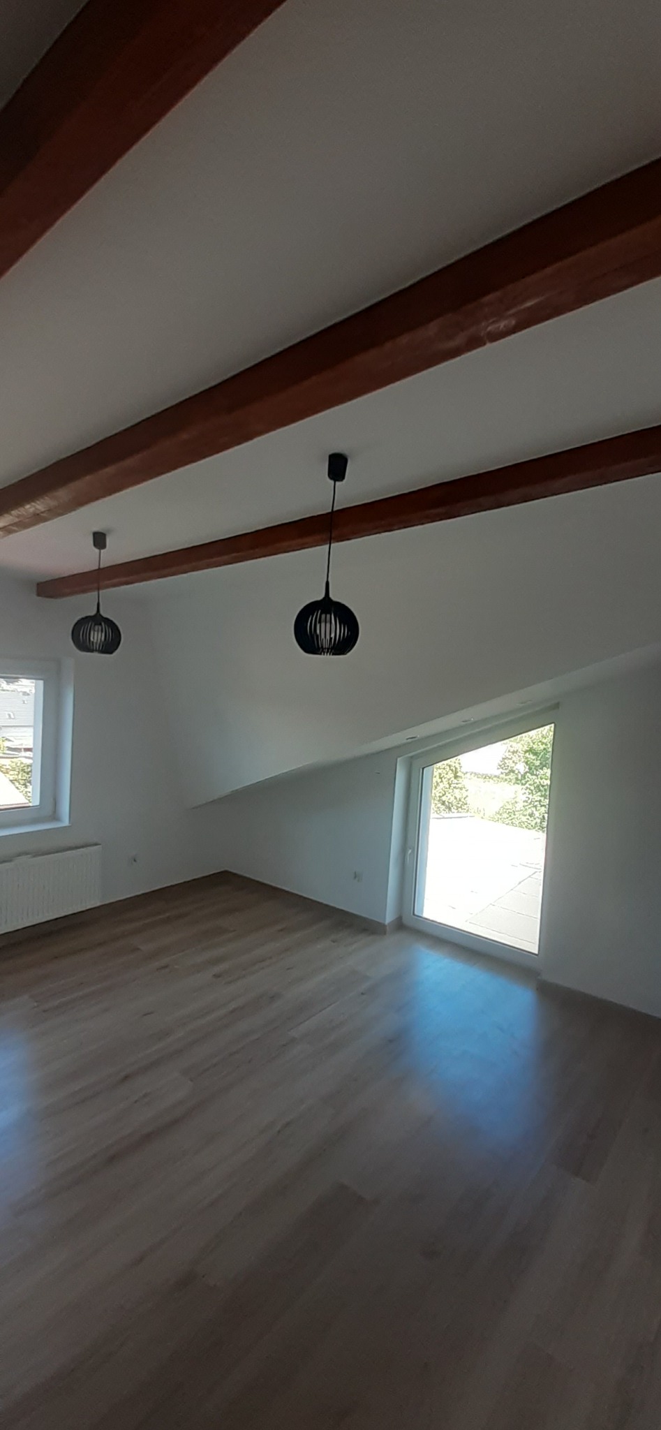 Poddasze po remoncie z drewnianymi belkami stropowymi, parkietem i nowoczesnym oświetleniem. Widoczne okno dachowe i standardowe okno z grzejnikiem.