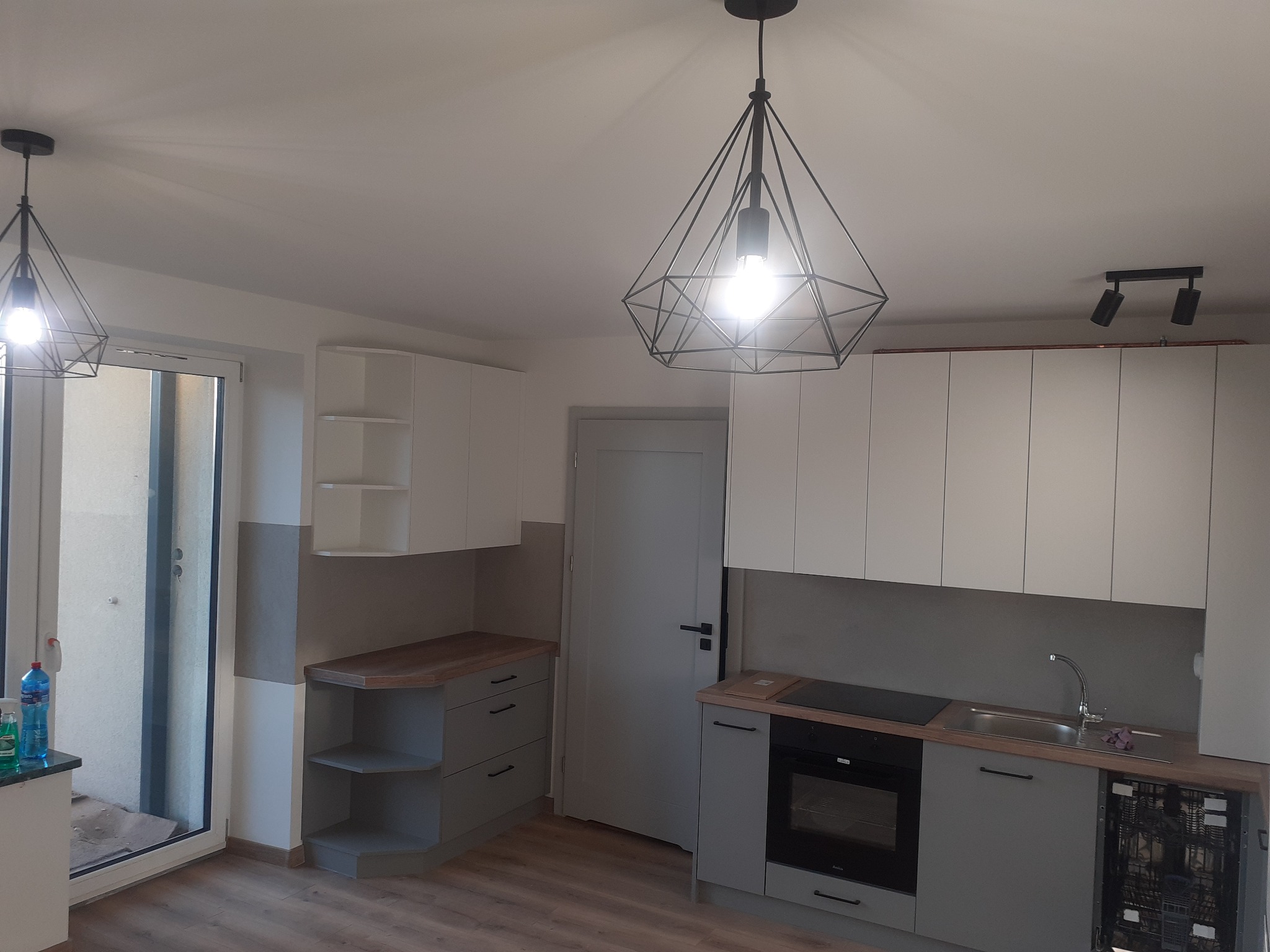 Nowoczesna kuchnia w szaro-białej kolorystyce z drewnianym blatem, oświetlona wiszącymi lampami w kształcie diamentów i dodatkowymi reflektorami sufitowymi, widok na otwarte drzwi balkonowe.