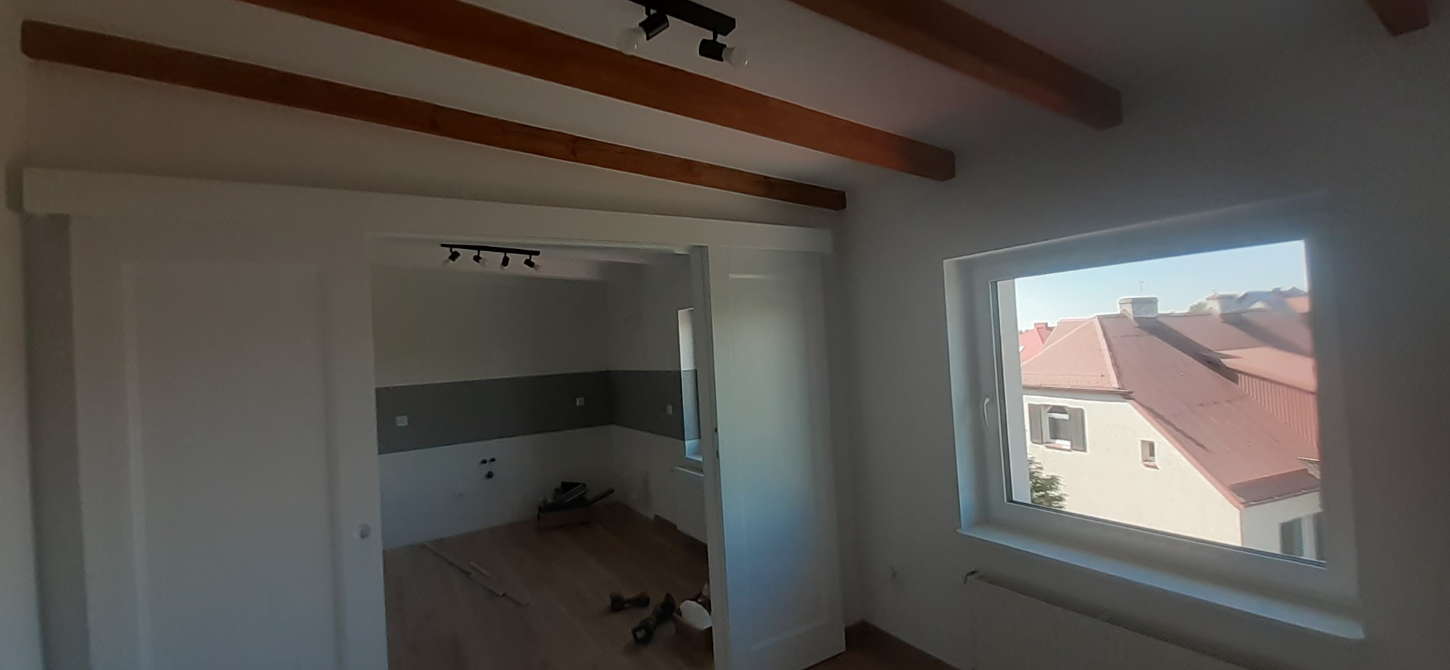 Wnętrze pokoju w trakcie prac wykończeniowych: położona drewniana podłoga, pomalowane ściany z szarym pasem, widoczne narzędzia i lampy sufitowe, okno z widokiem na budynek z czerwoną dachówką.