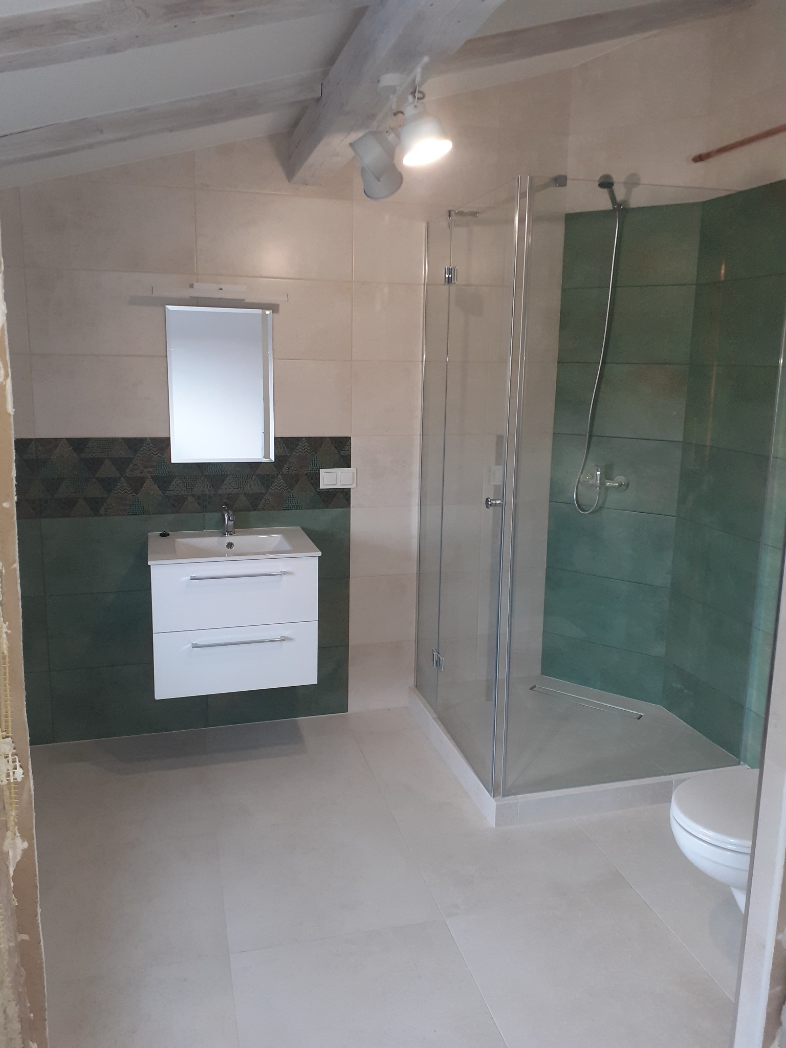 Nowoczesna łazienka na poddaszu z kabiną prysznicową, umywalką z szafką i zielonymi płytkami, wykończona w jasnych kolorach.