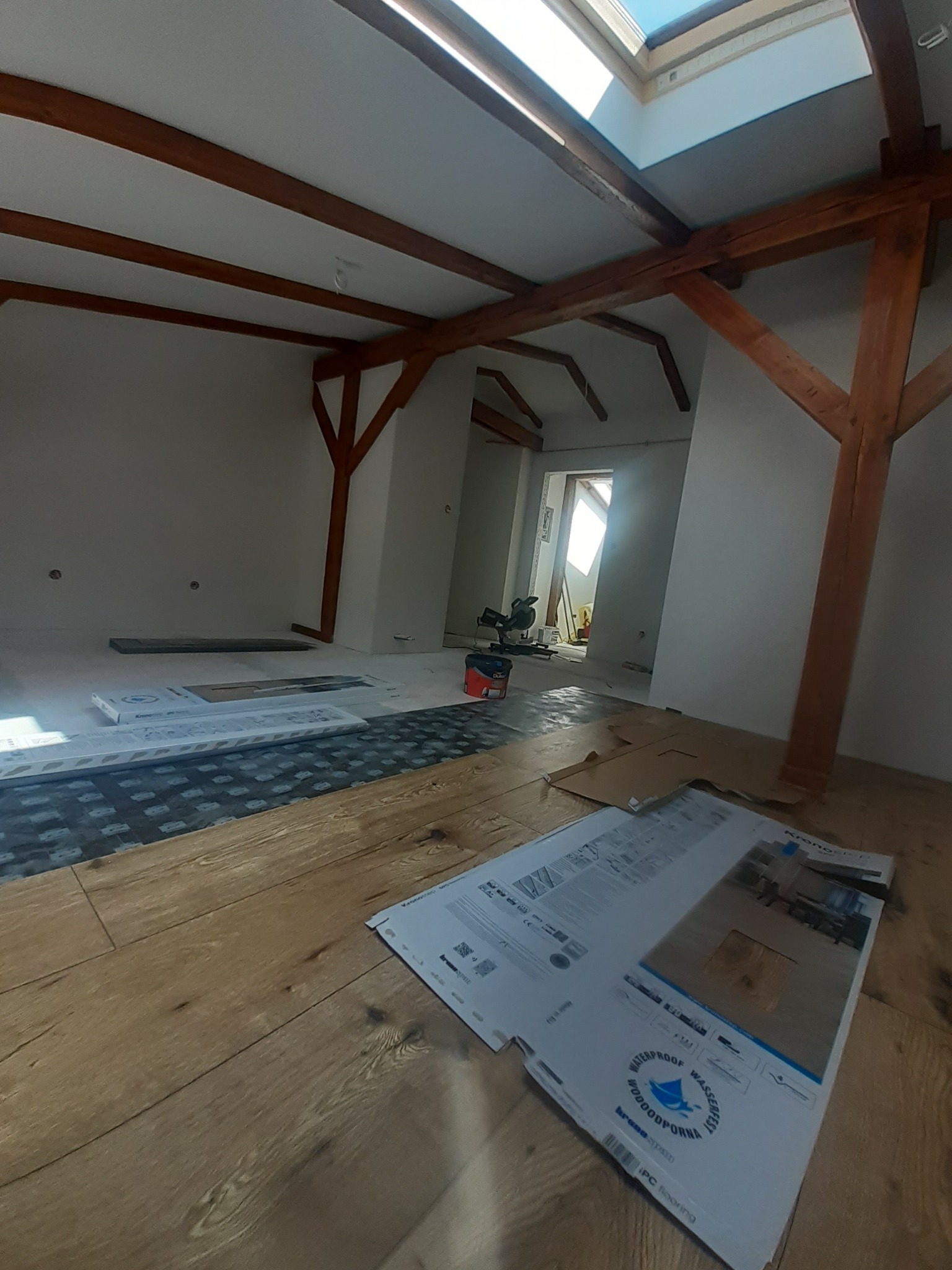 Poddasze w trakcie prac wykończeniowych: widoczne drewniane belki stropowe, okno dachowe, panele podłogowe, podkład wygłuszający i kartony z próbkami podłogi marki Kronopol.