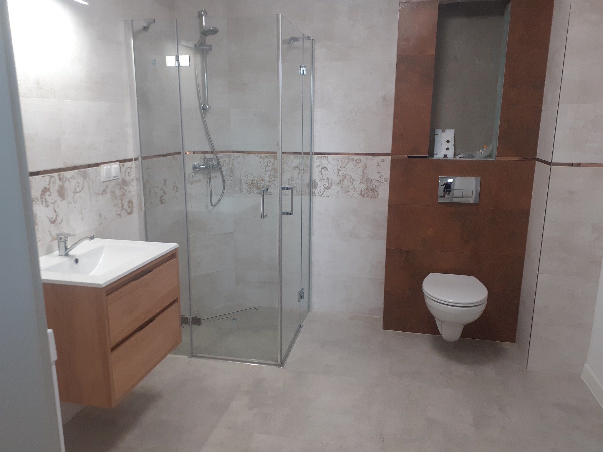 Nowoczesna łazienka po remoncie: kabina prysznicowa ze szklanymi drzwiami, umywalka z drewnianą szafką, podwieszana toaleta na tle brązowych płytek z wnęką.