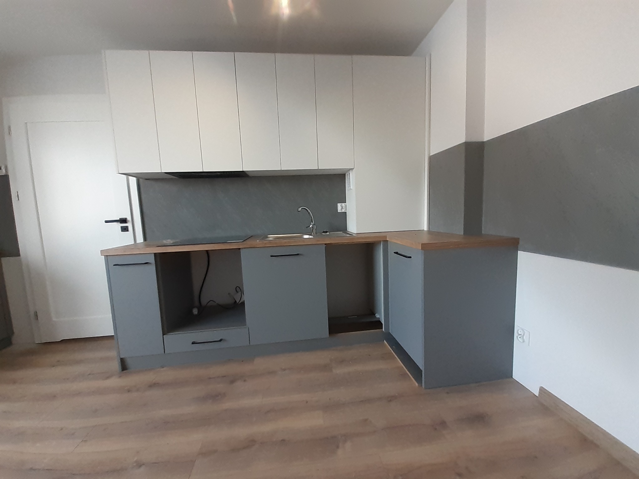 Nowoczesna kuchnia z szarymi szafkami dolnymi i białymi górnymi, drewniany blat, szara ściana nad blatem, parkiet na podłodze, widok na gniazdko elektryczne przy podłodze.