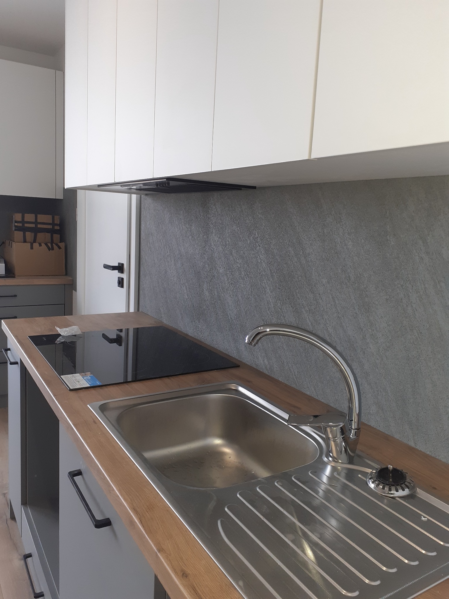 Nowoczesna kuchnia z drewnianym blatem, stalowym zlewem, płytą indukcyjną i białymi szafkami wiszącymi na tle szarej ściany.