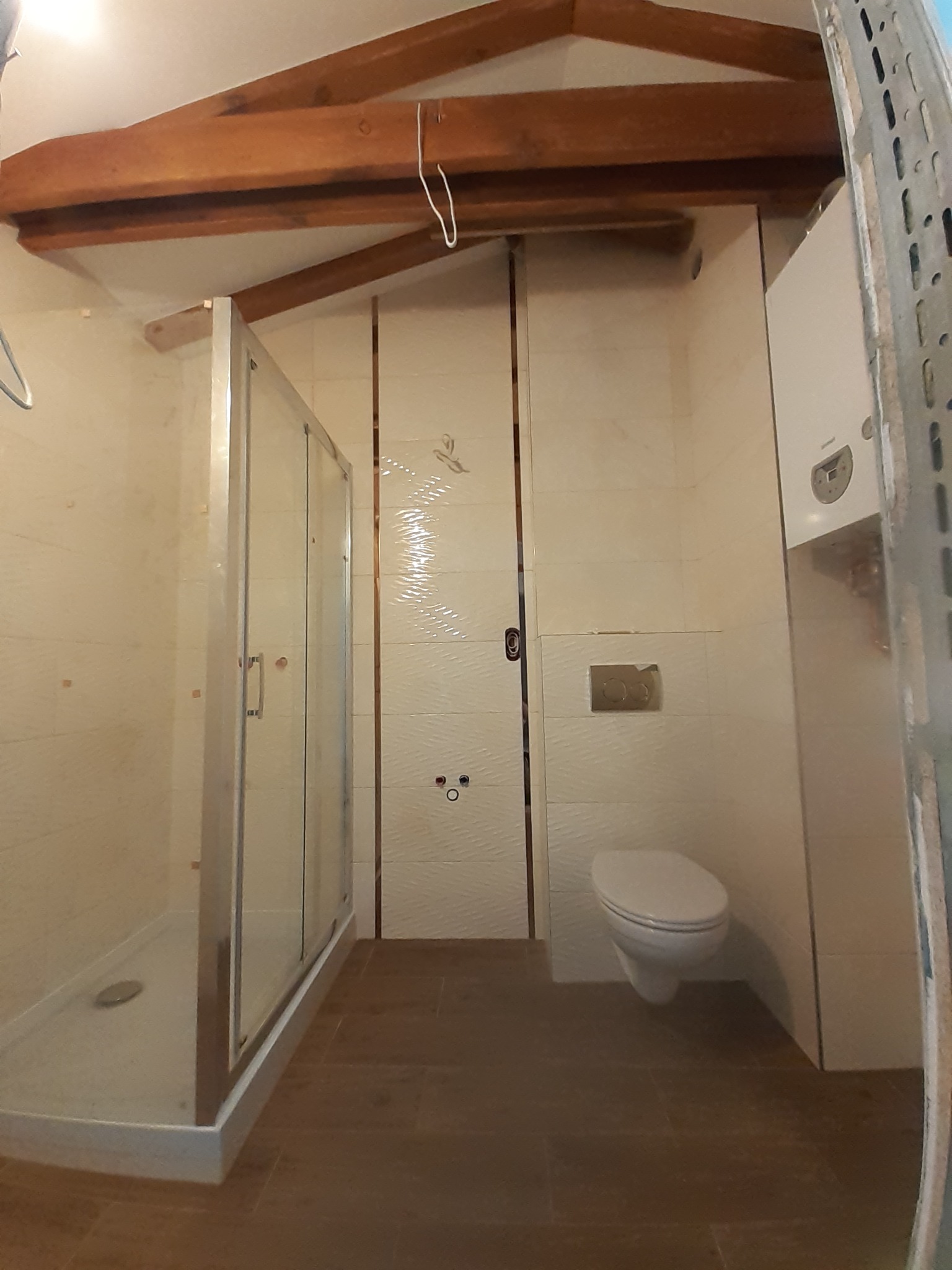 Nowa kabina prysznicowa ze srebrnymi profilami i brodzikiem, biała toaleta wisząca oraz piec gazowy w wykończonej łazience na poddaszu ze skośnym sufitem i drewnianymi belkami.