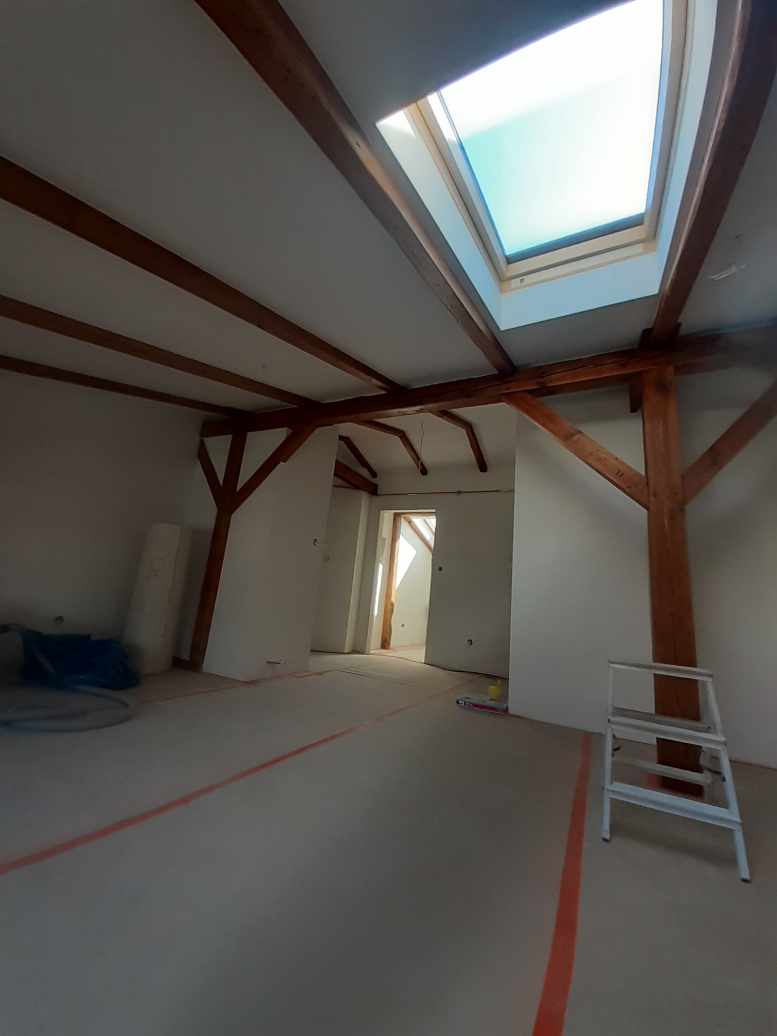 Poddasze w trakcie prac wykończeniowych: widoczne drewniane belki stropowe, okno dachowe, ślady po wyznaczaniu linii na podłodze oraz drabina.