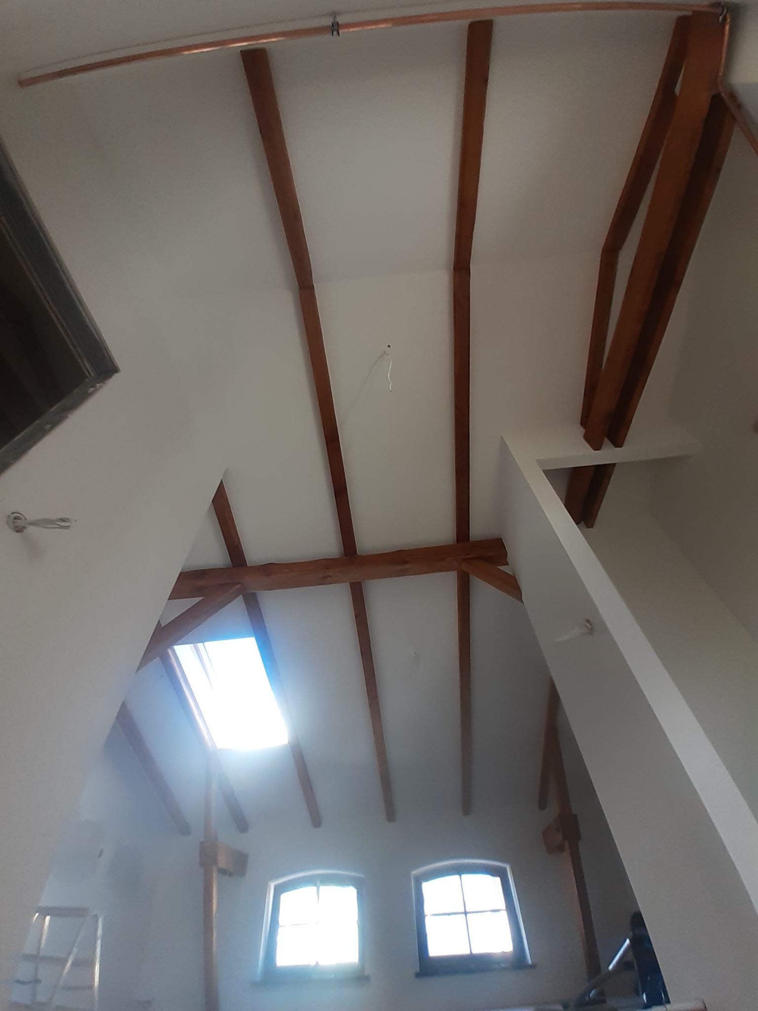 Poddasze z drewnianą konstrukcją więźby dachowej po malowaniu na biało, widoczne okna dachowe i pionowe, miedziana rurka pod sufitem.